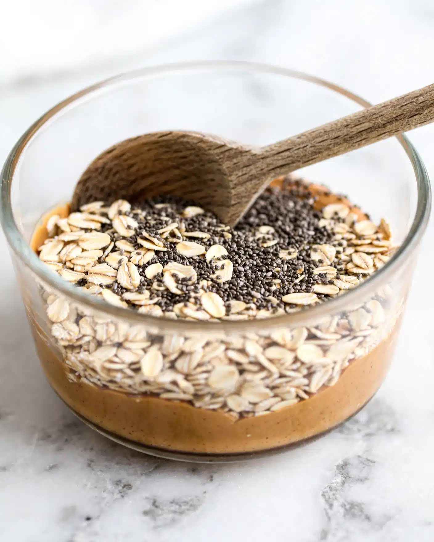Healthy No-Bake Breakfast Cookies Recipe 4 A clear glass bowl sits on a white marbled surface, holding three visible layers of ingredients: the bottom layer is smooth and light brown peanut butter, topped partly by a layer of rolled oats which are beige and flat, and a layer of small, round black chia seeds covering part of the oats. A wooden spoon rests inside the bowl, its round scoop touching the side with the peanut butter and oats. Photo taken with an iphone --ar 4:5 --v 7