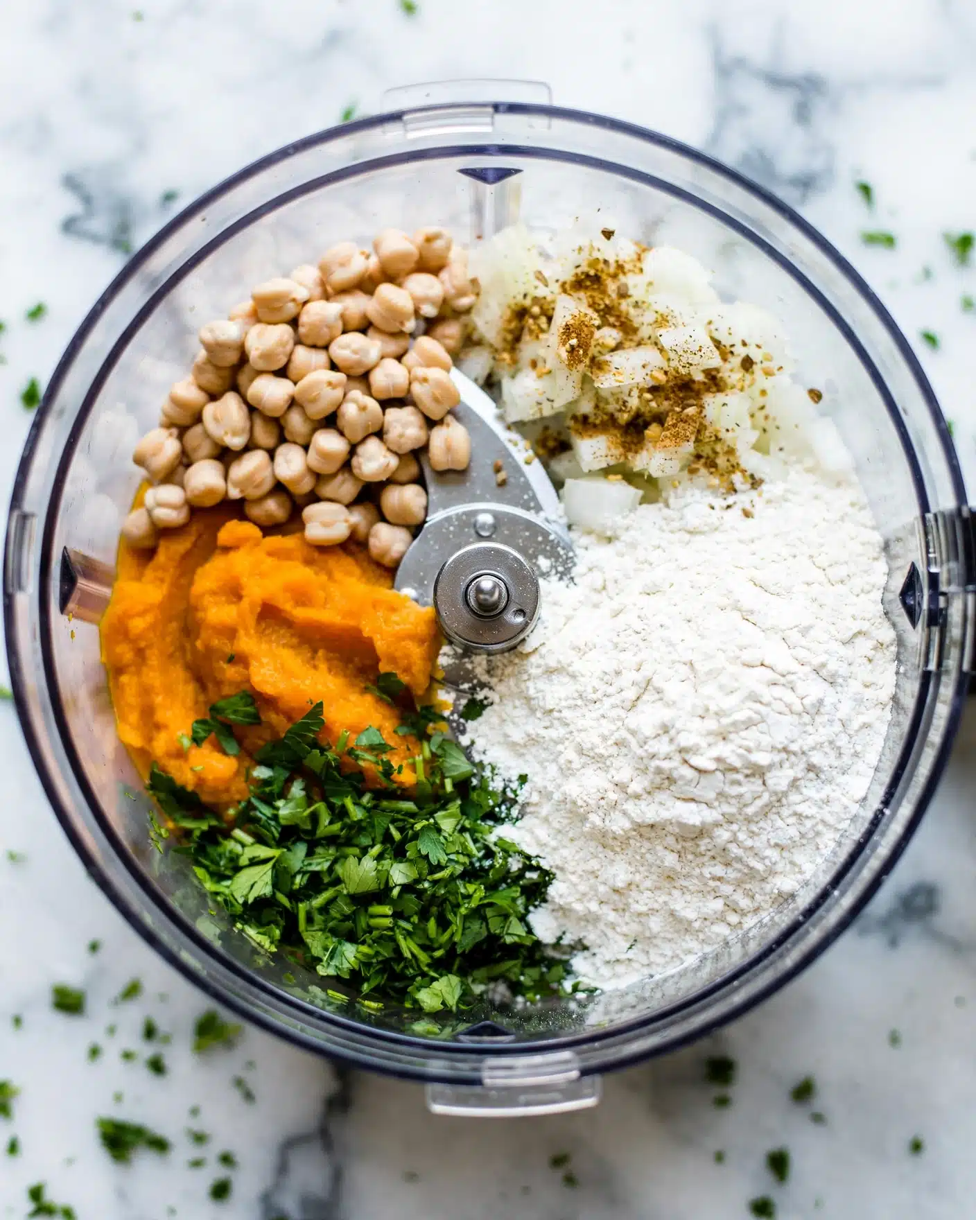 The image shows a food processor bowl filled with different ingredients before mixing. The bowl contains a white flour layer on the right side with a handful of chopped green herbs on the bottom left. Above the herbs is a bright orange mashed pumpkin layer. At the top left, there are white chickpeas, and on the top right, there are brown and light brown spices sprinkled over chopped white onions. The base of the bowl and the blades are visible in the center. The bowl is placed on a white marbled surface. photo taken with an iphone --ar 4:5 --v 7