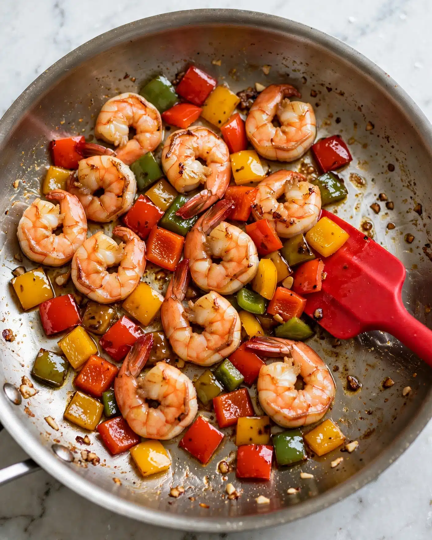The dish shows a close-up of cooked shrimp and colorful diced bell peppers in a pan. The shrimp are pink with light brown grilled marks and are mixed with red, yellow, and green bell pepper pieces that have a shiny, soft texture. The pan has a light coating of oil and small browned bits, and a red silicone spatula is visible stirring the mixture. The background is a white marbled texture. photo taken with an iphone --ar 4:5 --v 7
