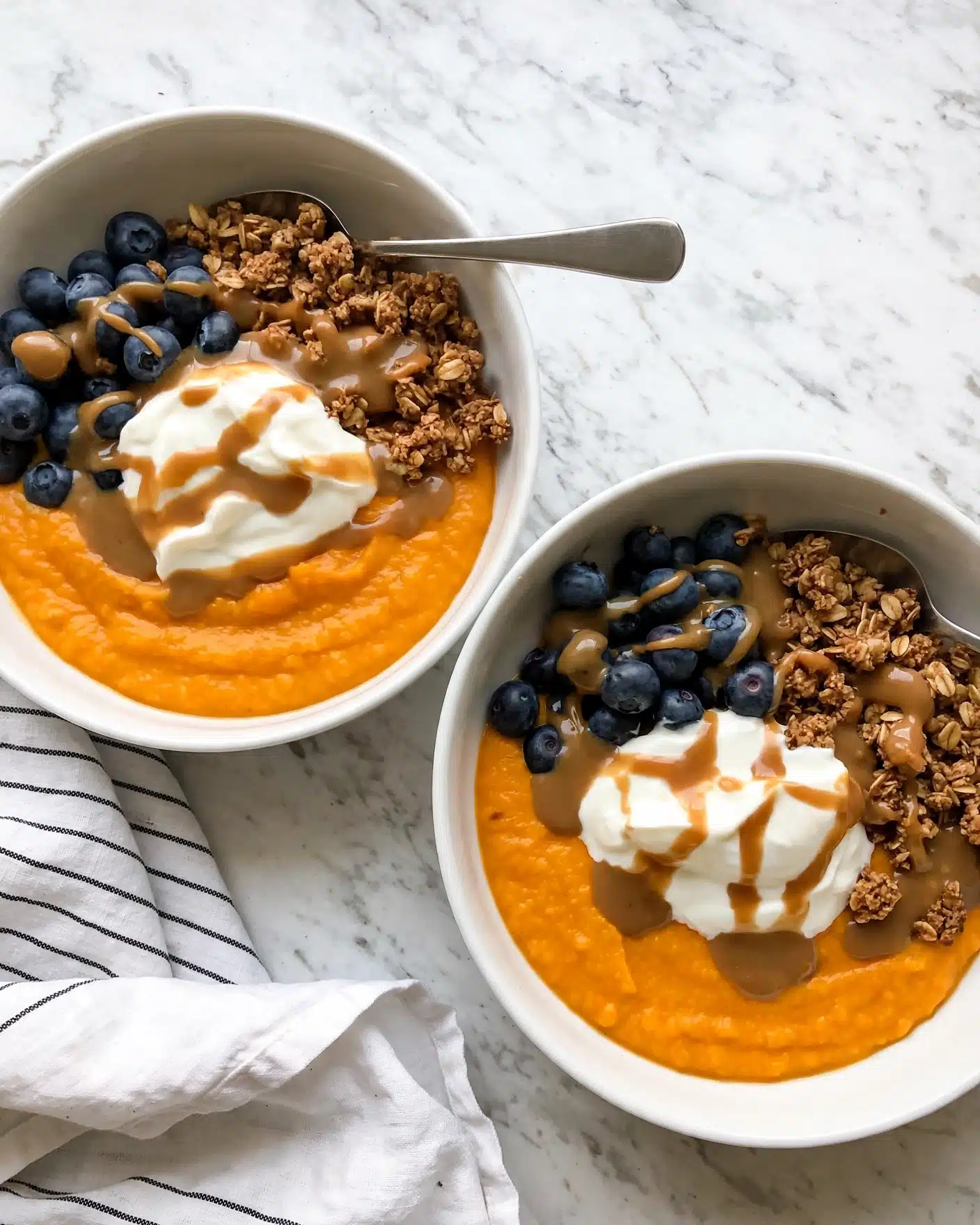 Two white bowls filled with three main layers are shown on a white marbled surface. The bottom layer is a smooth, bright orange puree covering most of the bowl. On one side, there is a layer of small, fresh, round dark blue blueberries. Next to the blueberries, a layer of crumbly, brown granola adds texture. On top of the orange puree and in the center, there is a dollop of creamy white yogurt with a drizzle of light brown sauce that also covers parts of the blueberries and granola. Each bowl has a silver spoon resting on the edge. A white cloth with thin black stripes is partially visible beside the bowls. Photo taken with an iphone --ar 4:5 --v 7