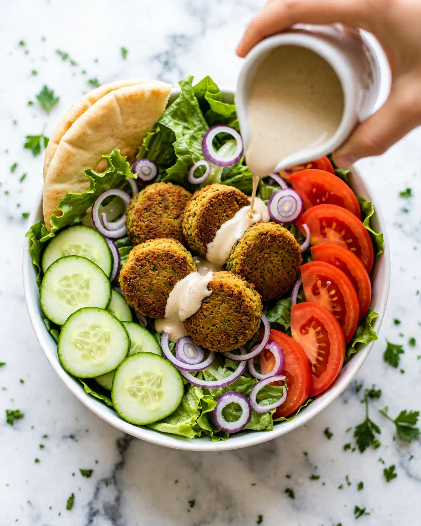A white bowl filled with a fresh salad that has many layers, starting with a base of green lettuce leaves, then topped with cucumber slices on the left side, bright red tomato wedges on the right, and thin rings of purple onion scattered around. In the center, there are five golden-brown falafel patties with a crispy texture. A white small jug is held by a woman's hand at the top right, pouring creamy beige tahini sauce over the falafel. There is a piece of pita bread tucked inside the bowl on the upper left side. The bowl sits on a surface with a white marbled texture, and some green herbs are scattered around for garnish. Photo taken with an iphone --ar 4:5 --v 7