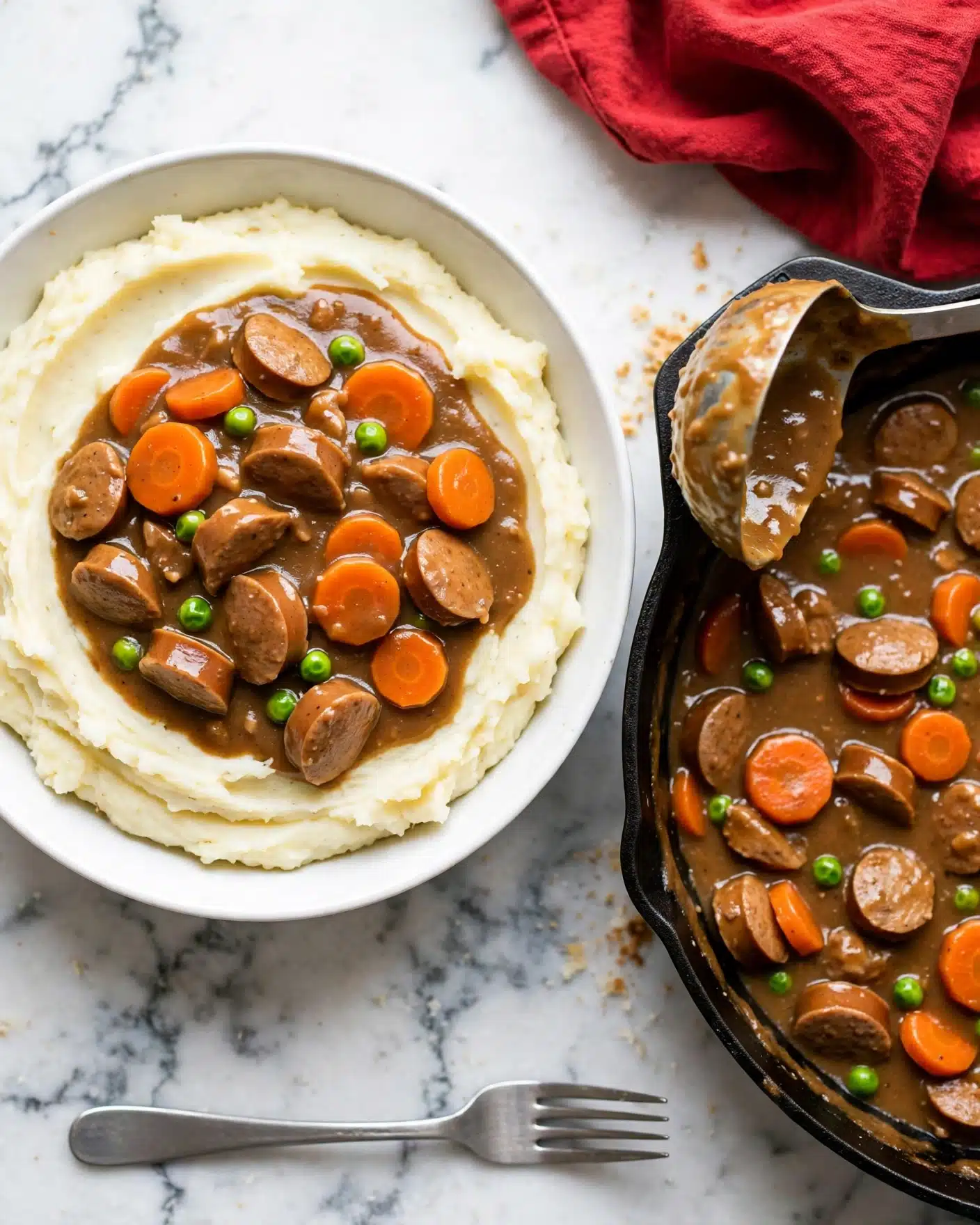 Curried Sausages Recipe 5 The image shows a white bowl filled with mashed potatoes forming the bottom layer, smooth and creamy with a light pale color. On top, there is a layer of brown stew with thick gravy, containing chunks of sausage-like pieces, bright orange carrot slices, and small green peas scattered throughout. Next to the bowl, there is a black pan with the same stew mixture inside, and a large silver ladle resting on the pan edge, partly covered with the brown sauce. The scene is set on a white marbled surface with a red cloth visible in the upper right corner, and a silver fork placed near the bottom of the image. photo taken with an iphone --ar 4:5 --v 7