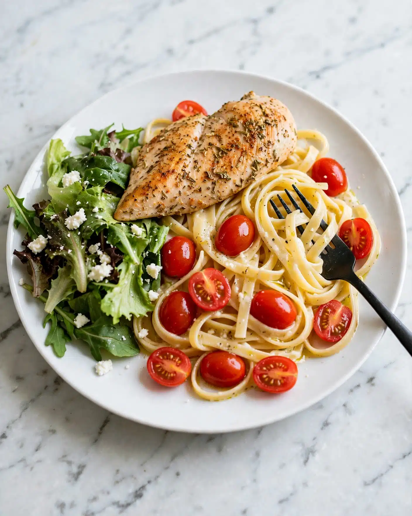 Creamy Pesto Chicken Recipe 5 A white plate with a piece of light brown seasoned chicken placed on top of a bed of pale yellow fettuccine pasta mixed with whole and halved bright red cherry tomatoes and some sauce. On the left side of the plate, there is a small pile of fresh mixed green leafy salad with dark and light green leaves and bits of white cheese scattered on top. A black fork rests on the right side of the plate, partially on the pasta. The background is a white marbled surface. photo taken with an iphone --ar 4:5 --v 7