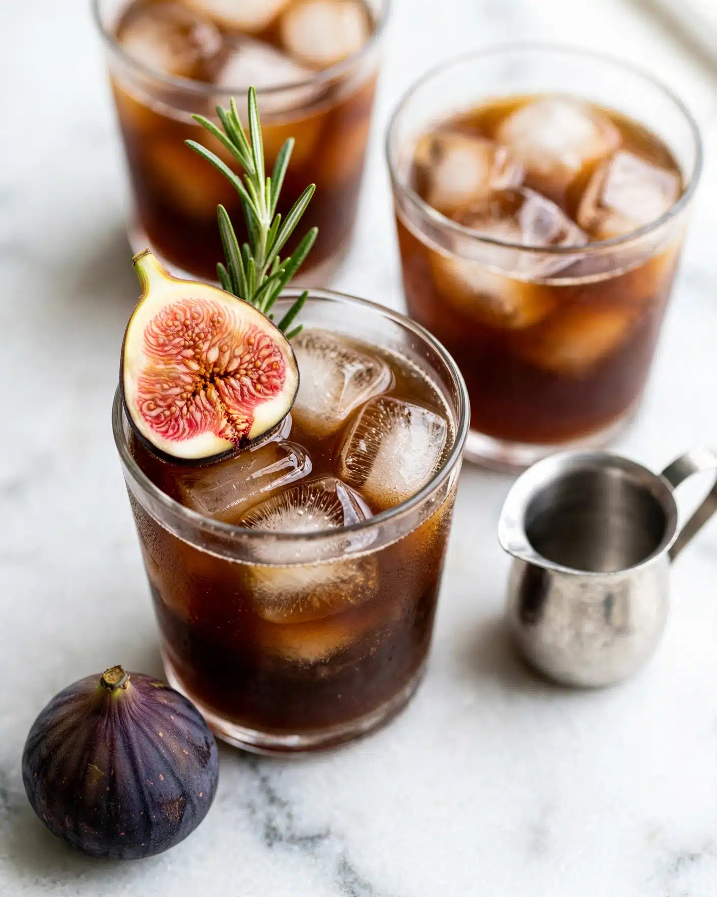A clear glass filled with a dark brown iced drink, layered with large ice cubes inside, topped with a fresh half fig showing its pink and seedy inside, along with a small green rosemary sprig standing upright next to the fig on the surface. The glass is placed on a white marbled surface with two similar glasses of the same drink and ice partially visible in the background. A whole fig with a dark purple skin lies beside the main glass, and a small silver pitcher is seen on the white marbled surface. Photo taken with an iphone --ar 4:5 --v 7