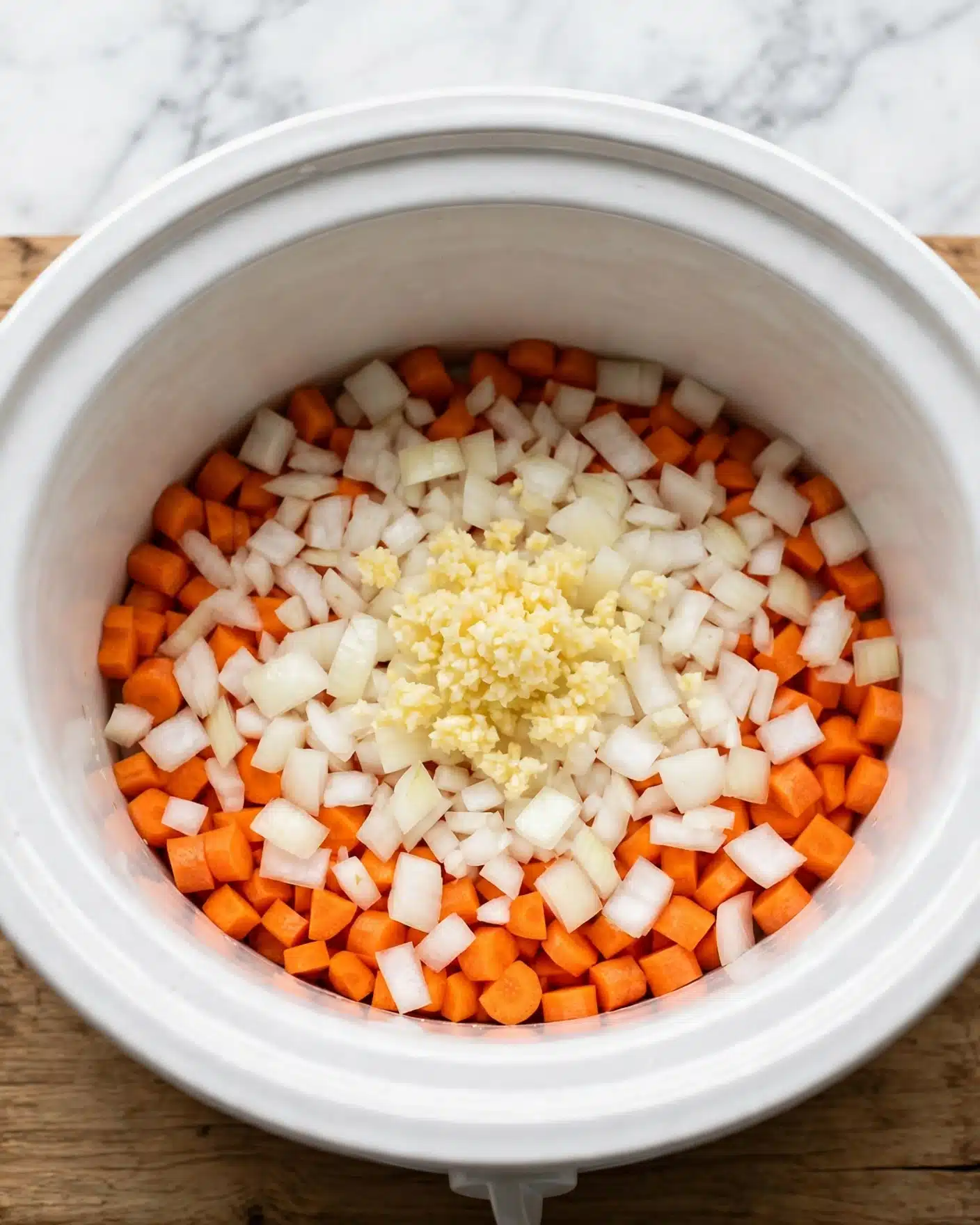 Easy Slow Cooker Cassoulet with Chicken and Sausage Recipe 4 Inside a white slow cooker pot, there is one layer of chopped orange carrots spread around the bottom, mixed with a layer of diced white onions unevenly covering the carrots, and a small pile of minced light yellow garlic sitting in the center on top. The pot is placed on a wooden surface, but the background should be imagined as a white marbled texture. photo taken with an iphone --ar 4:5 --v 7