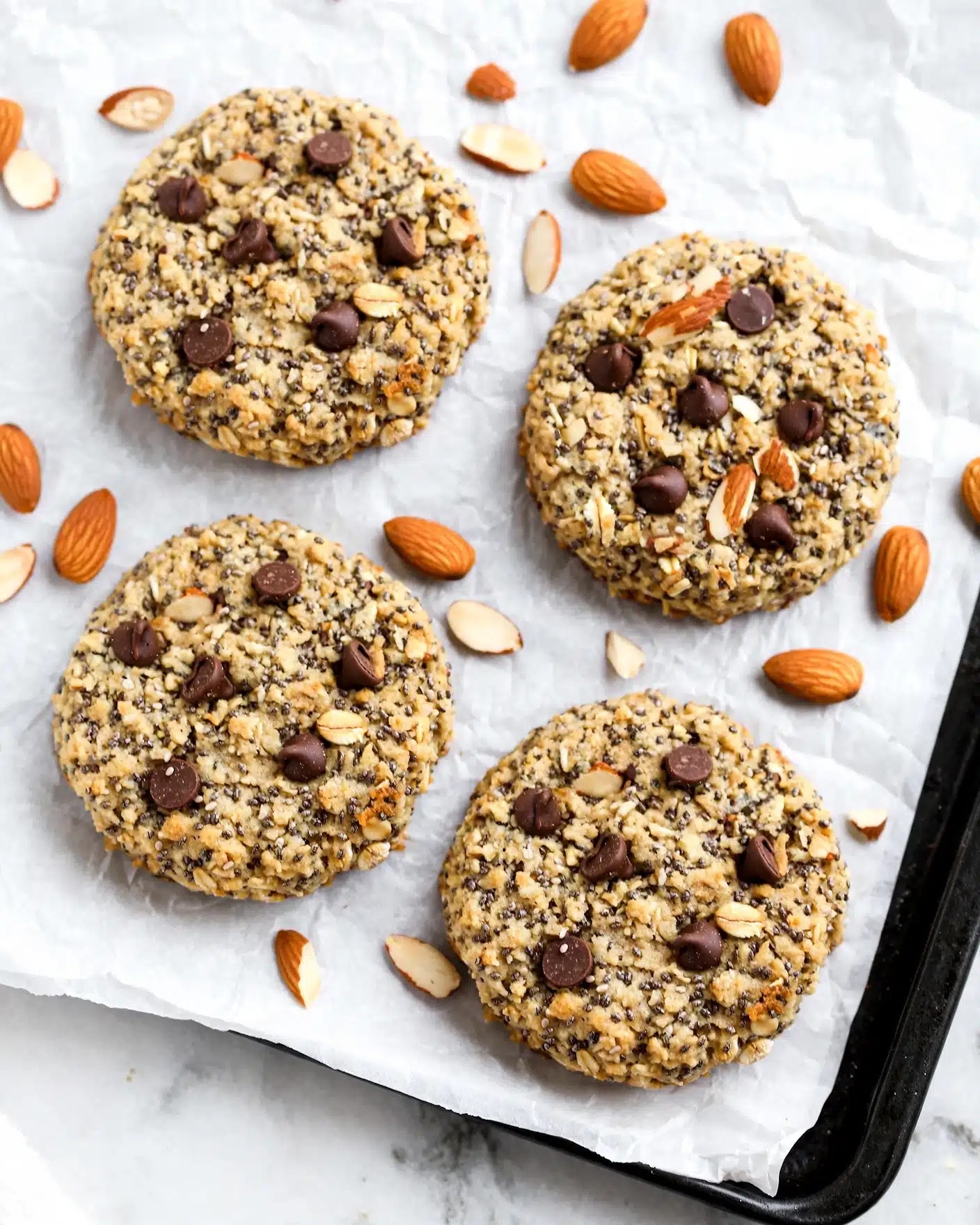 Healthy No-Bake Breakfast Cookies Recipe 5 Four round cookies with a golden-brown color, each filled with visible dark chocolate chips, chia seeds, oats, and pieces of almonds, laid out on a sheet of white parchment paper on a white marbled surface. The cookies have a rough texture with a mix of small and large bits of ingredients showing on the surface. Scattered whole and half almonds surround the cookies. The corner of the black baking tray holding the parchment paper is visible in the lower right. photo taken with an iphone --ar 4:5 --v 7