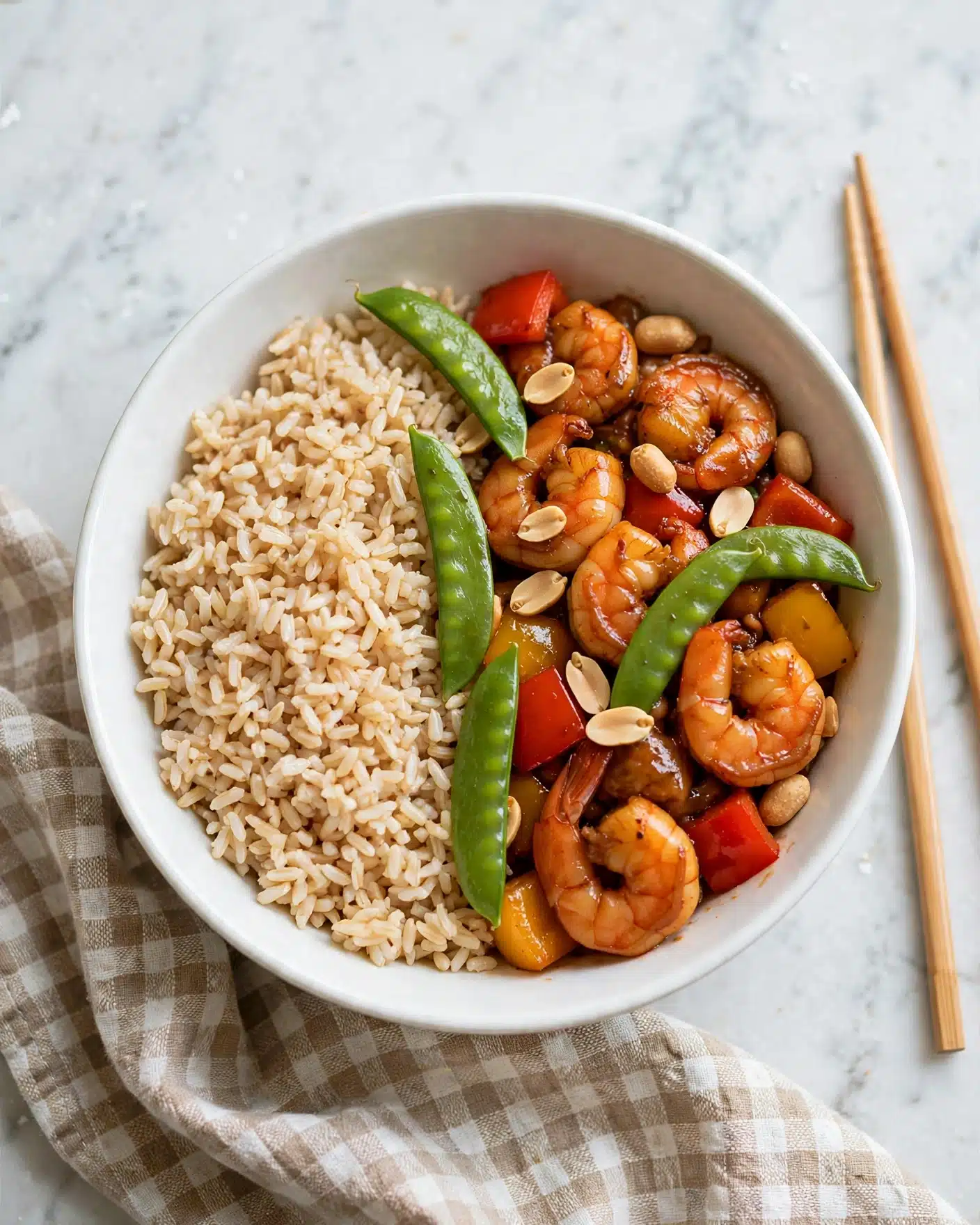 A white bowl filled with three main layers: on the left, a layer of cooked brown rice with a light, grainy texture in beige color; the right and center layers show orange-brown glazed shrimp, yellow and red bell pepper pieces, and green snap peas mixed with roasted peanuts scattered on top, all coated with a shiny sauce giving a glossy look. The dish is seen on a white marbled surface with a pair of chopsticks resting beside the bowl. Photo taken with an iphone --ar 4:5 --v 7