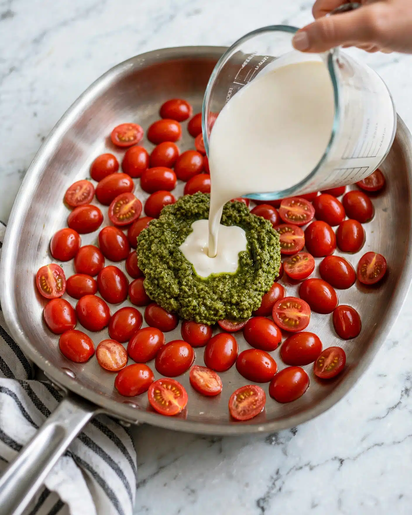 Creamy Pesto Chicken Recipe 4 In a silver metal pan on a white marbled surface, there is a layer of bright red cherry tomatoes, some whole and some cut in half, spread across the pan. On top of the tomatoes, there is a medium-sized dollop of green pesto sauce with a textured, leafy look. A woman's hand is pouring a smooth, thick white cream into the pan from a transparent glass measuring cup, creating a contrast with the red tomatoes and green pesto. The image shows the moment just before the cream mixes with the other ingredients. Photo taken with an iphone --ar 4:5 --v 7