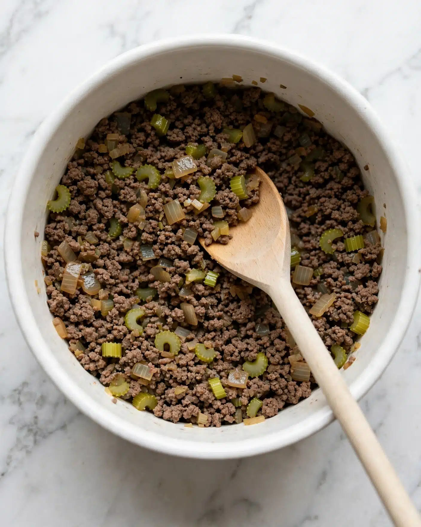 Inside a white pot, there is a mix of cooked ground meat with small, soft, light green celery pieces and translucent light brown onion bits. The meat is dark brown and crumbly, spread evenly across the bottom. A light wooden spoon lies on top of the mixture, resting diagonally from the bottom right toward the center. The pot sits on a white marbled surface. photo taken with an iphone --ar 4:5 --v 7
