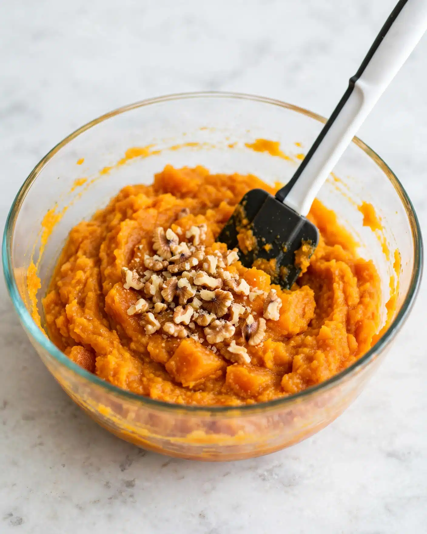 Sweet Potato Breakfast Parfait Recipe 4 A clear glass bowl with a thick orange mixture inside, resembling mashed pumpkin or sweet potato. On top of the orange base is a pile of small broken walnut pieces, slightly off-white with brown edges, sitting in the center. A spatula with a white handle and black rubber head is placed inside the bowl, touching both the walnuts and orange mix. The bowl rests on a white marbled surface photo taken with an iphone --ar 4:5 --v 7