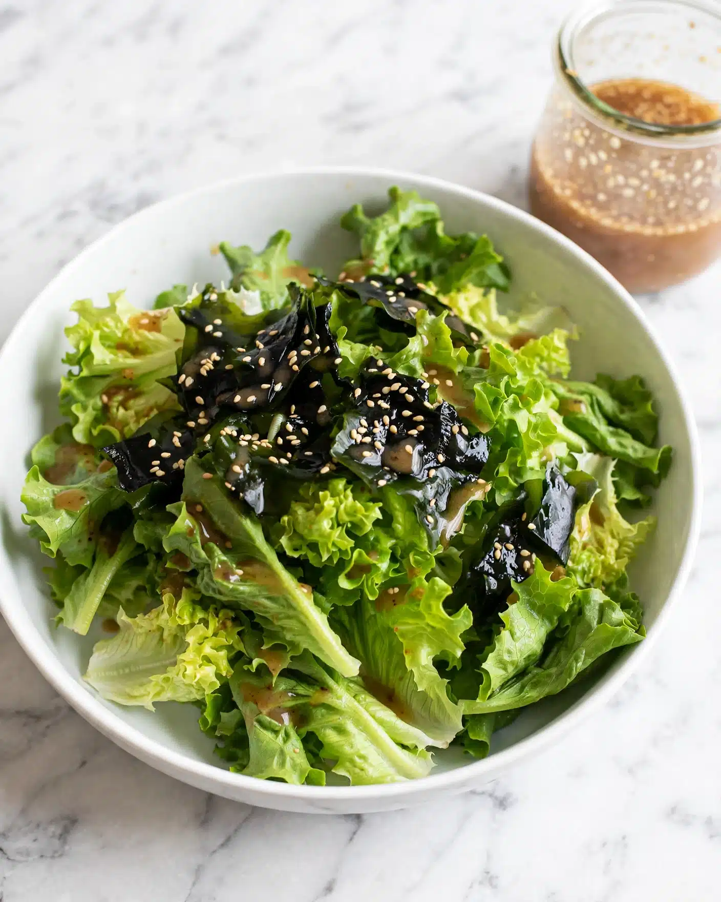 Easy Wakame Salad (Japanese Seaweed Salad) Recipe 5 A white bowl filled with a fresh salad showing two main layers; the bottom layer is made of light green lettuce leaves with a soft and crisp texture, while the top layer consists of shiny dark green seaweed pieces sprinkled with small sesame seeds. A light brown dressing is gently drizzled over the layers. The bowl is placed on a white marbled surface, and in the background to the right, there is a glass jar containing extra dressing. photo taken with an iphone --ar 4:5 --v 7