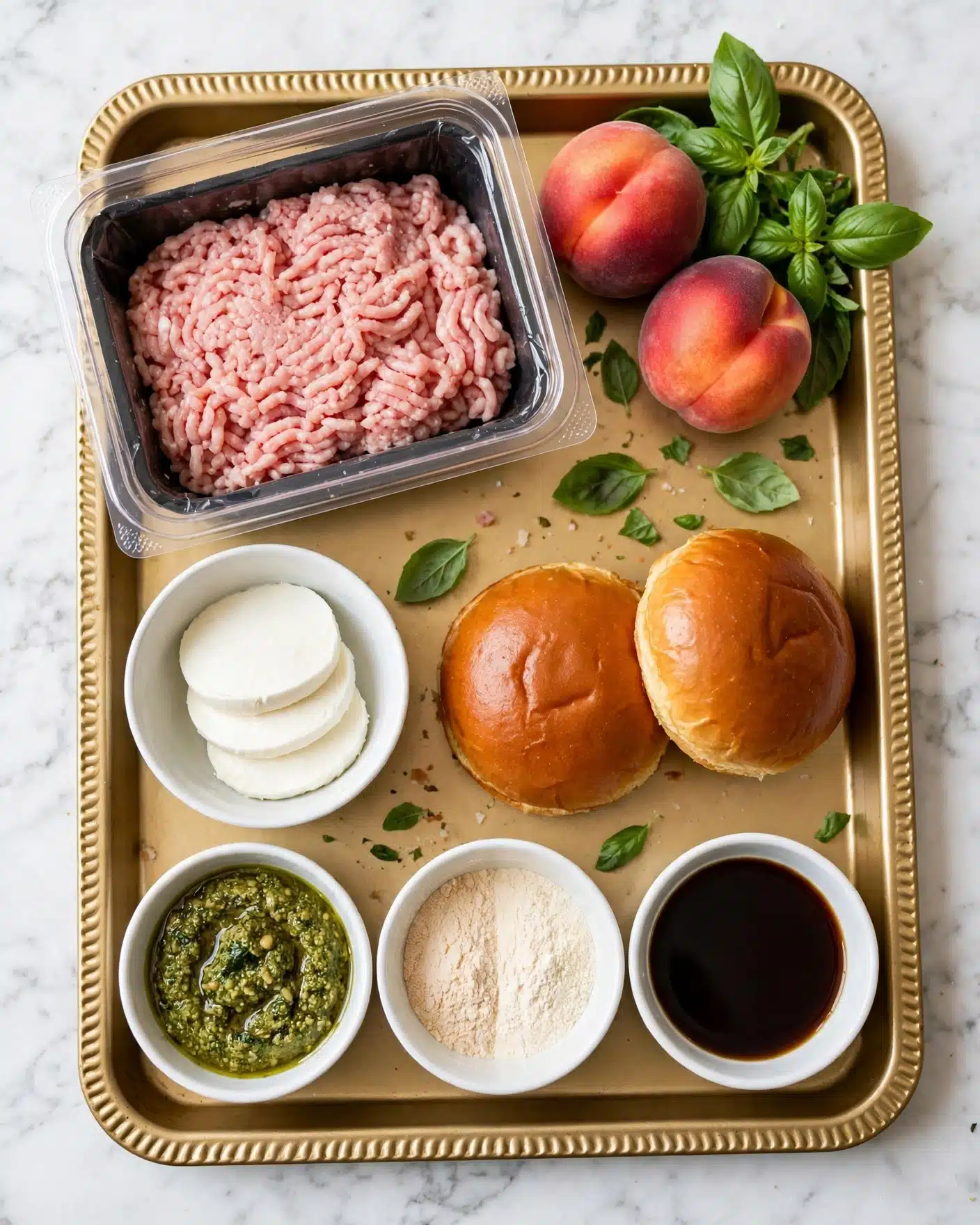 Peach Caprese Burgers Recipe 4 The image shows a gold baking tray on a white marbled surface holding several ingredients: at the top left, a clear plastic container with pale pink ground meat; to the right, two reddish peaches and fresh green basil leaves arranged together; below the meat, a small white bowl with three smooth white mozzarella slices; next to the mozzarella, two shiny golden brown burger buns side by side; below the buns, three small white bowls, the left one filled with a green pesto sauce showing texture, the middle one holding two different light powders, and the right one with a dark brown liquid resembling soy sauce. Photo taken with an iphone --ar 4:5 --v 7