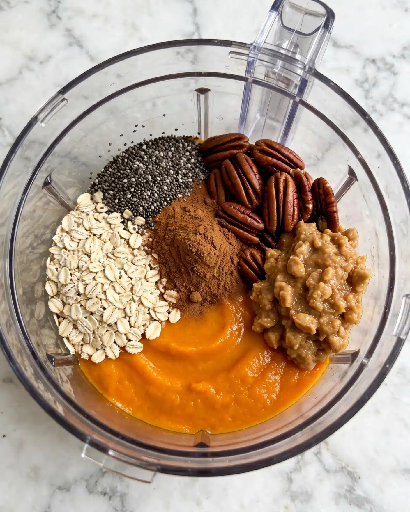 A clear food processor bowl with six distinct ingredients arranged in layers: light beige rolled oats on the left, black and white chia seeds above them, a rough brown powder of cinnamon in the center, glossy dark brown pecans to the right, a chunky light brown paste-like layer beside the pecans, and a bright orange smooth pumpkin puree at the bottom, all set on a white marbled surface photo taken with an iphone --ar 4:5 --v 7