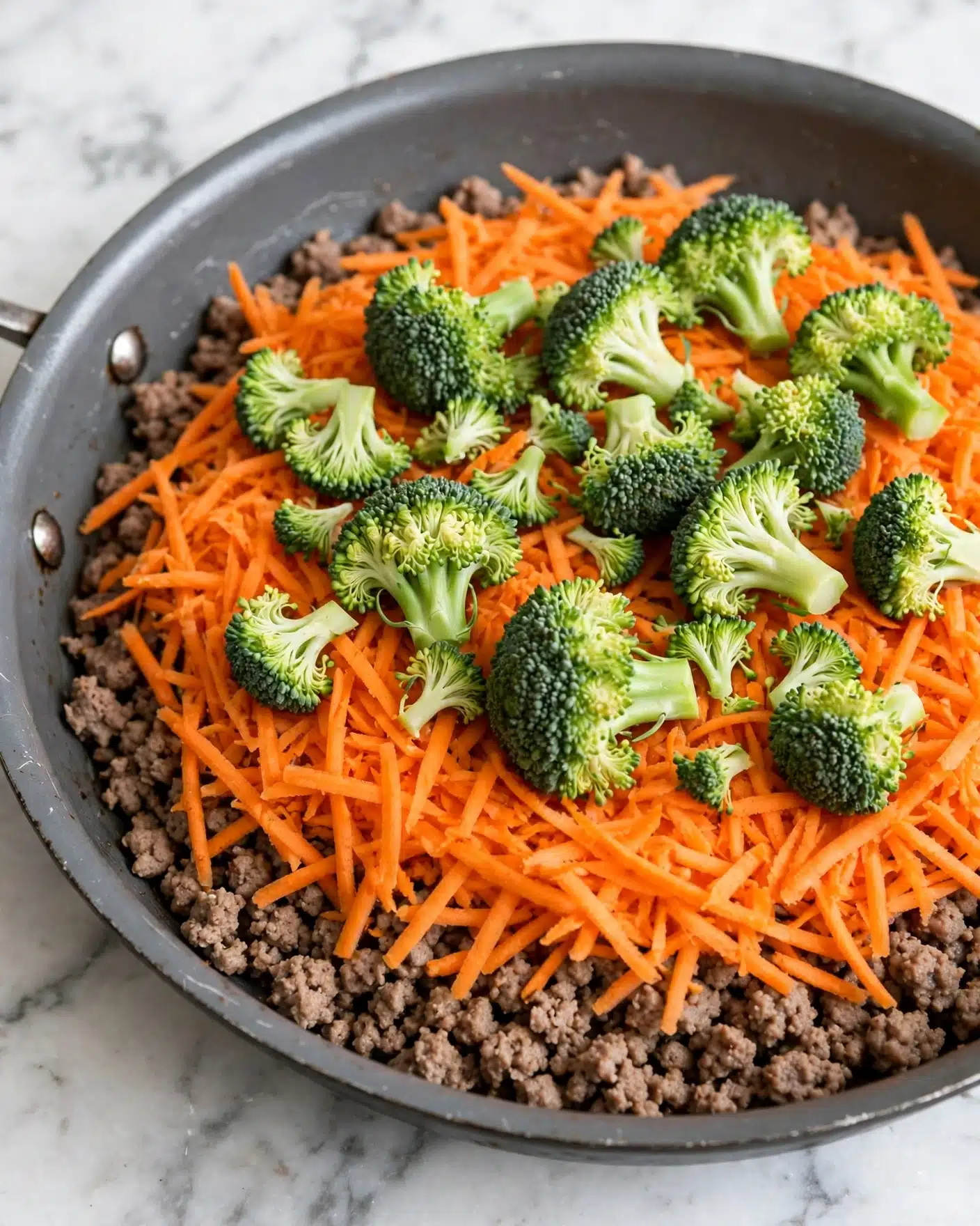The image shows a close-up of a gray cooking pan filled with three layers of food, placed on a white marbled surface. The bottom layer consists of a cooked ground meat base with a light brown color and crumbly texture. The middle layer is a thick sprinkling of bright orange shredded carrots spread evenly across the meat. The top layer is fresh, green broccoli florets with a rough, bumpy texture, placed loosely over the carrots. The mix of green and orange creates a fresh and vibrant look. photo taken with an iphone --ar 4:5 --v 7