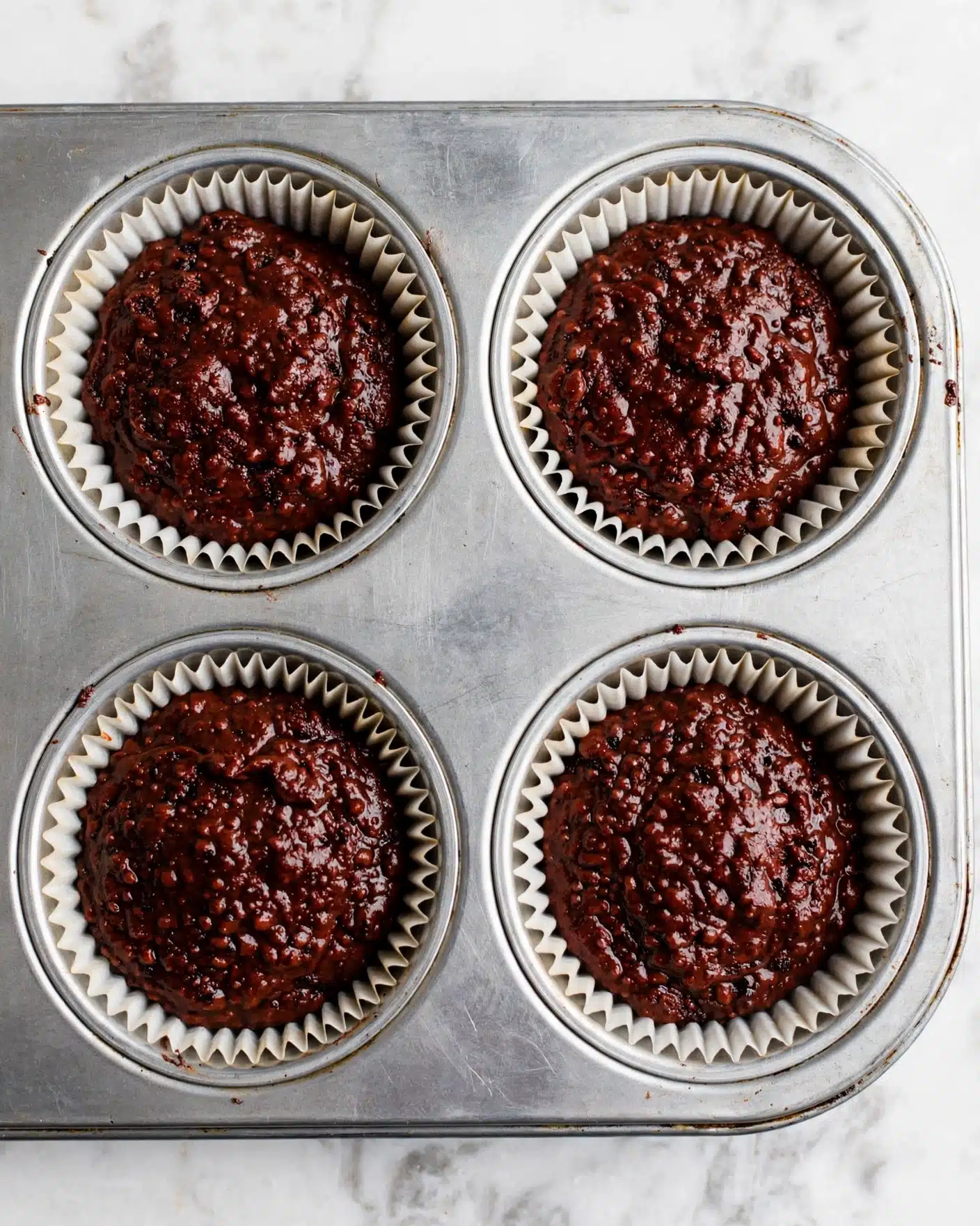 Double Chocolate Beet Cupcakes Recipe 4 The image shows a close-up of a metal cupcake tray with four round cavities, each filled with thick, dark red-brown batter that looks moist and textured. Each cavity holds a white paper cupcake liner, neatly folded around the edges, containing the rich batter nearly full to the top. The tray rests on a white marbled surface, creating a clean and simple background. photo taken with an iphone --ar 4:5 --v 7