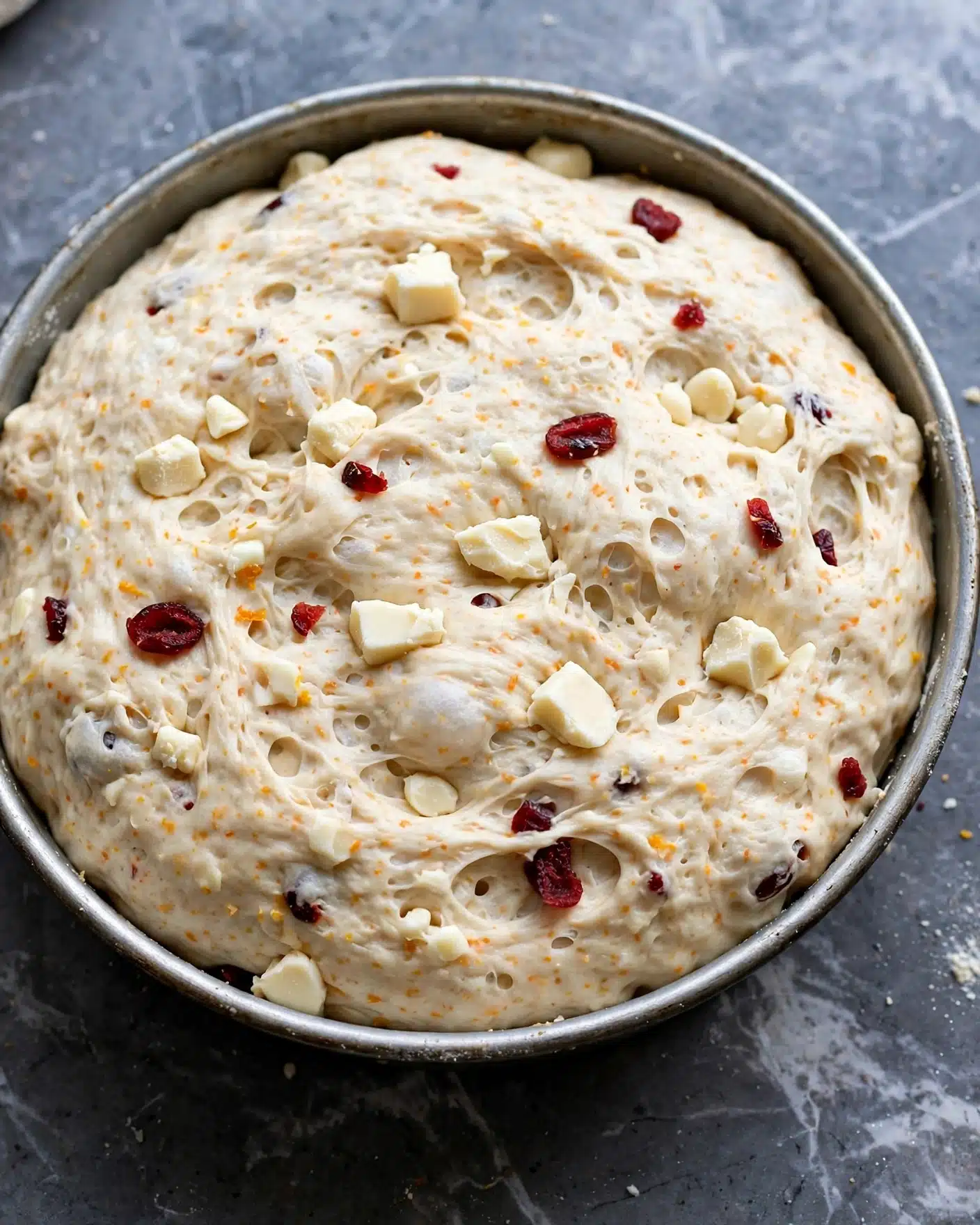 A close-up image of a round metal pan filled with a thick, pale dough that has risen and looks soft and fluffy. The dough has small visible pieces of white chunks and a few dried red fruit bits scattered on top and partly embedded inside. The dough surface is slightly uneven with some small holes, and there are faint orange specks throughout. The pan is placed on a dark gray surface with a white marbled texture background. photo taken with an iphone --ar 4:5 --v 7