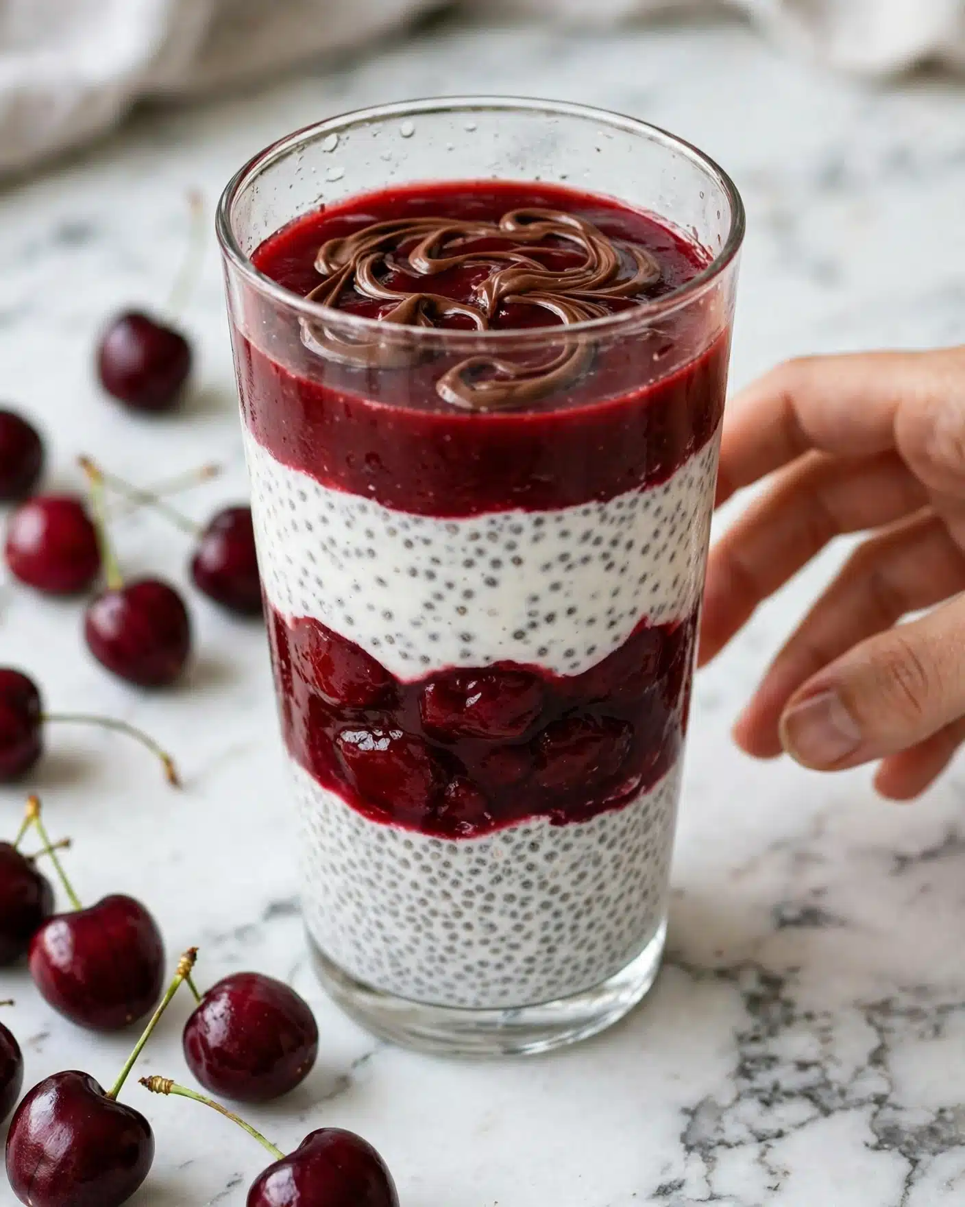Vegan Black Forest Chia Seed Pudding Parfait Recipe 5 The image shows a clear glass filled with four visible layers. The bottom layer is white with a speckled texture, looking like chia pudding. Above it is a thick, dark red cherry sauce layer. The third layer is again the white chia pudding, and the fourth layer is the dark red cherry sauce, visibly rich and smooth, topped with swirls of melted chocolate. Around the glass on a white marbled surface, dark red cherries with green stems are scattered, and a woman's hand is reaching from the right side of the frame. photo taken with an iphone --ar 4:5 --v 7