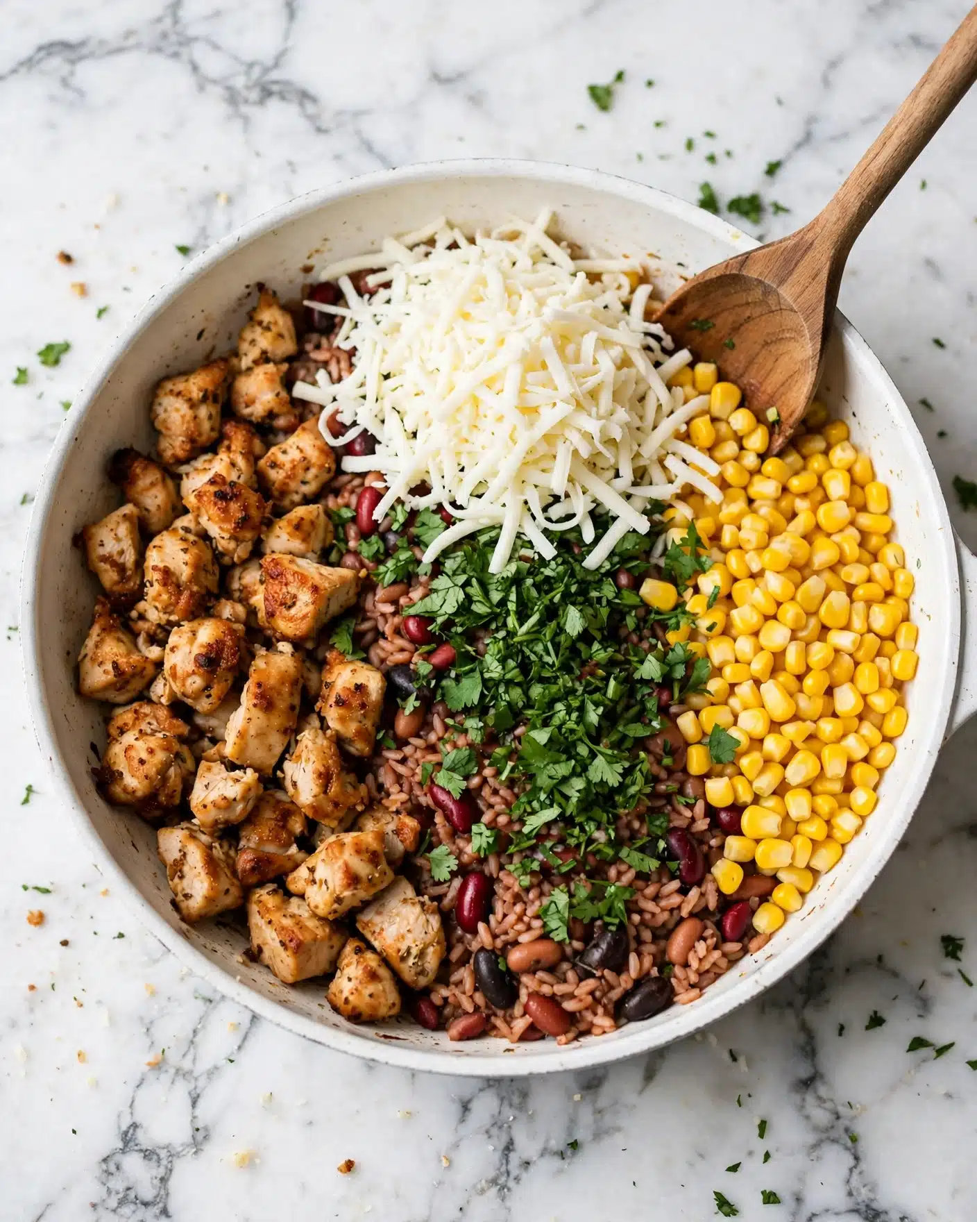 Cheesy Southwest Chicken and Rice Recipe 4 The image shows a white pan filled with cooked food divided into sections, placed on a white marbled surface. On the left, there is a layer of golden-brown cooked chicken pieces with a slightly crispy texture. In the middle, there is a pile of shredded white cheese sitting above some green chopped herbs and a mixture of red and brown cooked rice and beans. To the right, there is a bright yellow layer of cooked corn kernels. A wooden spoon rests on the pan's edge, partly inside the food. Photo taken with an iphone --ar 4:5 --v 7