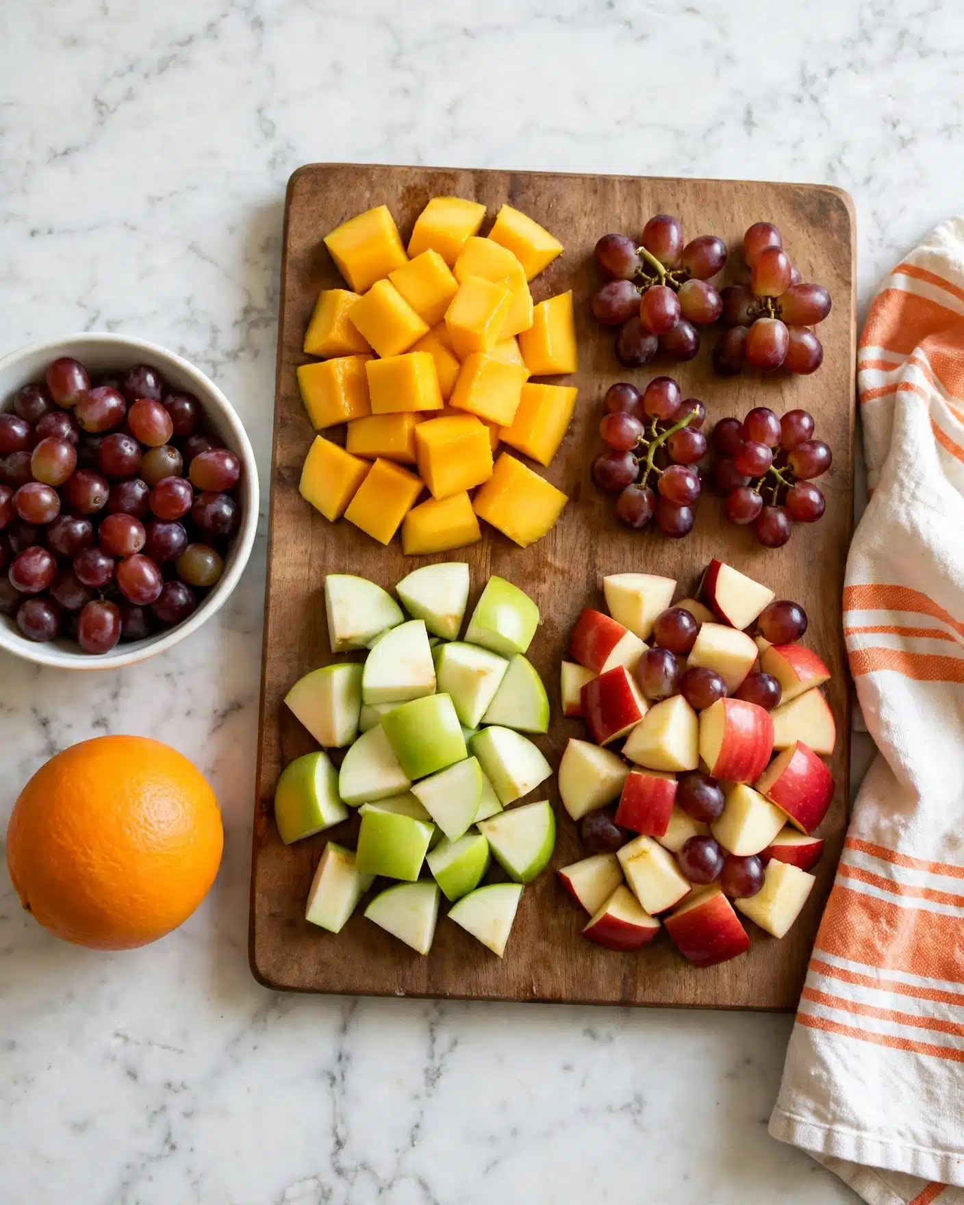 Fall Fruit Salad Recipe 4 A wooden board on a white marbled surface holds four types of chopped fruit neatly arranged in separate sections: small red grape clusters at the top right, bright yellow-orange fruit pieces at the top left, light green apple chunks at the bottom left, and red apple pieces mixed with red grapes at the bottom right. Next to the board, a white bowl filled with small red grapes is visible on the left, and a whole orange sits in the foreground to the left. On the right side of the board, a kitchen towel with orange stripes is partly visible. Photo taken with an iphone --ar 4:5 --v 7