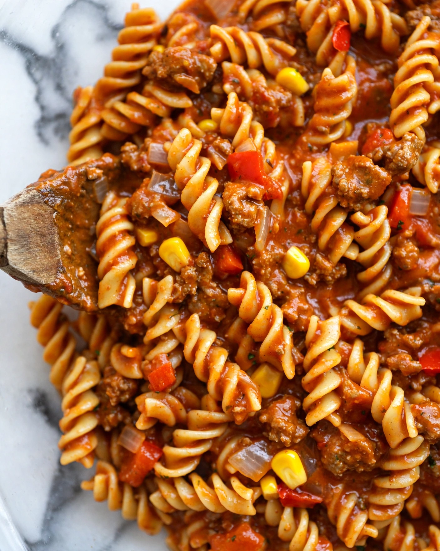 A close-up view of a cooked pasta dish with spiral pasta spirals covered in a thick reddish-orange sauce. Mixed within are small bits of browned ground meat, bright yellow corn kernels, and small pieces of red bell pepper along with finely chopped onions. The texture looks soft and saucy, with the ingredients well blended. A wooden spoon is partially visible stirring the mix against a white marbled background. Photo taken with an iphone --ar 4:5 --v 7